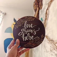 "Love grows here." This simple, elegant message is hand-painted on a round, dark-stained wooden plaque, perfect for celebrating a new home, a wedding, or the love in your family.