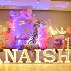 A full view of the 'Anaisha' first birthday stage with large, illuminated marquee letters. The butterfly theme with pastel balloons and mushroom props creates a fairytale atmosphere.