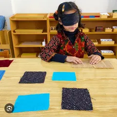A blindfolded child uses her sense of touch to match different fabric textures. This classic Montessori sensorial activity refines the tactile sense, builds vocabulary for description, and develops concentration.