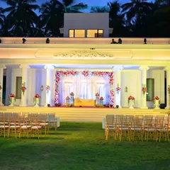 A grand, white colonial-style stage setup for an outdoor reception. The structure is adorned with beautiful pink and white floral arrangements, creating a classic and elegant look.