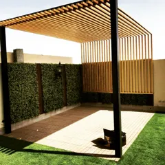 A modern pergola design combining a metal frame with a wooden slatted roof and side screen for partial privacy. The structure is set on a tiled patio next to an artificial turf lawn and a green wall.