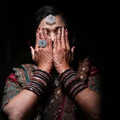 An artistic bridal portrait focusing on the intricate henna on the bride's hands as she covers her face, with dramatic lighting highlighting her jewelry.