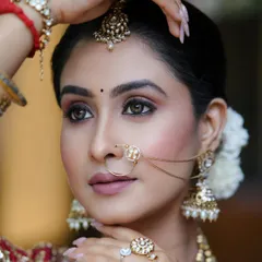 A stunning close-up of a bridal photoshoot look. The focus is on the intricate eye makeup and the flawless, glowing skin, complemented by traditional jewelry like the nath and maang tikka.