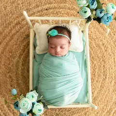 A newborn wrapped in mint green fabric sleeps in a miniature white crib, surrounded by matching blue flowers for a coordinated and peaceful scene.
