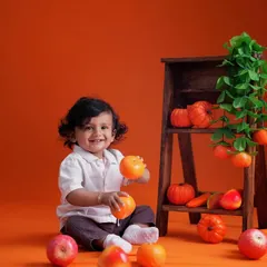A happy toddler sits amidst a vibrant orange-themed setup, playing with oranges and pumpkins against a matching backdrop.