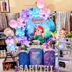 An enchanting 'under the sea' party with our Little Mermaid theme. This home setup for Sahithi's 5th birthday includes a custom backdrop with Ariel, a purple and teal balloon arch, and mermaid-themed cake plinths.