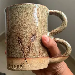 A beautiful stoneware mug with a unique double handle and delicate lavender sprigs carved into the clay. The speckled, sandy glaze gives it a rustic, natural charm.