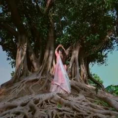 A powerful and artistic portrait of a woman in a pink lehenga, posing amidst the intricate roots of a massive banyan tree.
