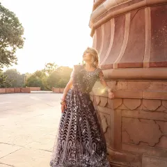 A full-length shot of the model leaning against a historic sandstone pillar. The warm sunlight and her upward gaze create a feeling of serenity and grace, showcasing the full splendor of the lehenga.