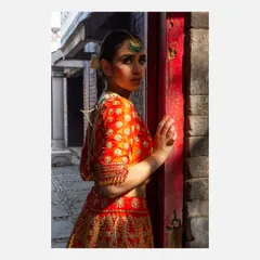 A striking portrait of a model in a traditional orange lehenga, looking over her shoulder in a narrow alleyway. The play of light and shadow emphasizes the rich color of the garment and the model's expression.