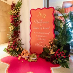 The welcome signage for the Mehendi celebration, themed 'Hues of Love'. The design featured a vibrant color palette of pink and orange, fresh fruits, and floral arrangements to greet guests.