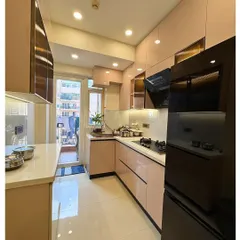 A beautiful and functional L-shaped modular kitchen in a soft pink hue. The design incorporates glossy cabinets, a white countertop, and a black chimney and fridge for a modern, contrasting look.