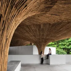 A quiet moment of conversation within the monumental bamboo structure of the TAPMI Centre. The scale of the columns creates intimate pockets within the larger communal space, fostering both public and private interactions.
