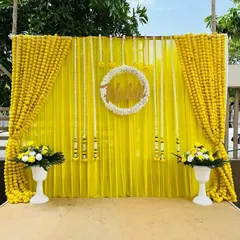 An elegant and bright Haldi backdrop created with yellow drapes, strings of white rajnigandha flowers, and a central floral wreath. The marigold borders and flower pots complete this classic look.