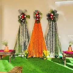 This is a modern and minimalist Haldi backdrop we created. The design uses three triangular frames with lush green turf as a base, featuring vibrant orange marigold curtains and white floral strings, topped with fresh flower bouquets for a clean and contemporary look.