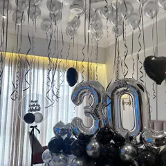 A chic and modern 30th birthday surprise decoration using a black and silver color scheme. The setup includes large silver '30' foil balloons, a bed arrangement, and matching balloons on the ceiling.