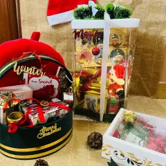 A festive collection of Christmas hampers. This display includes a large transparent bag with goodies, a round velvet 'Merry Christmas' box, and a smaller decorative gift box.