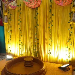 A simple and traditional Haldi backdrop for a home function. The setup includes a bright yellow curtain, decorative umbrellas, and a low wooden stool (chowki) for the ceremony.