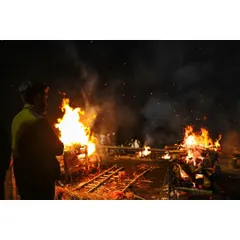 A man stands watching the intense flames of multiple funeral pyres at night. This image focuses on the raw power of the fire and the human element of observation and reflection.