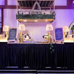 A stylishly decorated bar for a cocktail event. The setup features custom art-deco-inspired signage, elegant table lamps, and lush greenery, all set against a dark, sophisticated tablecloth.