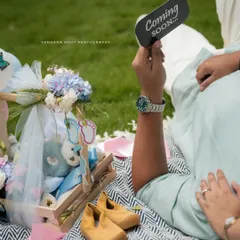 A close-up on the details of a maternity picnic, showing a "Coming Soon" sign, a gift basket, and tiny baby shoes.