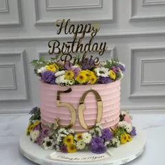 Celebrating a 50th birthday with a burst of color. This cake features a pink ribbed buttercream finish and is adorned with a crown and base of fresh, vibrant wildflowers in shades of purple, yellow, and white.