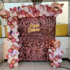 A terrace party with a rose gold sequin wall. The setup features a "Happy Birthday" neon sign and a balloon arch in shades of pink and white with butterfly accents.
