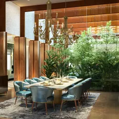 A formal dining room where a large, traditional chandelier hangs over a table surrounded by plush velvet chairs. A wall of glass opens the space to an indoor courtyard, seamlessly blending biophilic design with classic luxury.