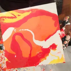 A close-up of a freshly poured canvas, showcasing a warm palette of red, orange, and yellow. The fluid motion of the paint creates stunning, organic shapes.