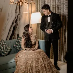 A full-length portrait showcasing the couple's impeccable style. The bride, seated in her magnificent golden lehenga, looks up at her groom, who stands beside her in a sharp tuxedo, creating a look of regal elegance.