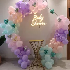 A chic and simple baby shower decoration. This setup features a circular metal frame adorned with a pastel rainbow balloon garland and a 'Baby Shower' neon sign, with a small table ready for a cake or gifts.