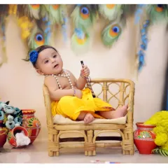 This baby is posed as Krishna, sitting in a miniature wicker chair and holding a flute. The setup includes cotton-filled matkas and a backdrop of blue curtains and peacock feathers.