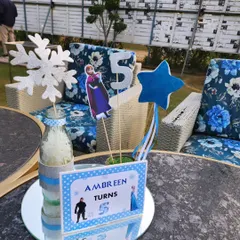 A Frozen-themed table centerpiece. It features cutouts of Anna and a sparkling number '5', along with a glittery snowflake, all set on a mirrored base to enhance the icy feel.