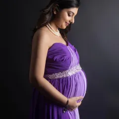 A profile shot capturing the beautiful curve of the baby bump. The mom-to-be wears a strapless purple gown with an embellished waistline, her gentle smile reflecting a moment of quiet joy.