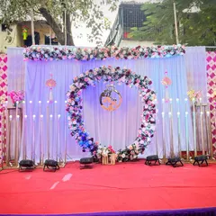 A lovely outdoor engagement setup on a red carpet. The centerpiece is a large floral ring decorated with pink and white flowers, set against a white curtain backdrop and flanked by modern, tall candle-like lights.