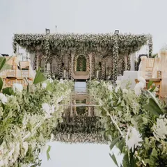A view down the stunning mirrored aisle leading to the all-white floral stage. Lined with lush arrangements of white flowers and greenery, the reflective surface creates a breathtaking effect, doubling the beauty of the decor. This is how I create a grand and dramatic entrance for the couple.