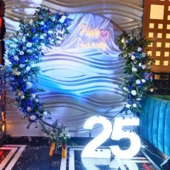 This 25th anniversary setup has a cool and elegant vibe. I used a floral arch with blue and white flowers, a neon sign, and large light-up "25" numbers against a modern, textured wall.
