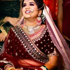The pure joy of a bride on her wedding day. This candid photo captures a moment of genuine laughter, showcasing her beautiful smile. The warm lighting enhances the rich maroon color of her velvet lehenga and the sparkle of her elaborate pink and white jewelry set.