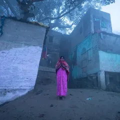 A woman in a bright pink saree stands alone in the dense morning fog at Yamuna ghat, a striking splash of color in a muted, mysterious landscape.