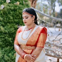 A serene portrait of a South Indian bride on her wedding day. The soft, glowing makeup and classic red lip perfectly match the elegance of her off-white and red Kanchipuram saree and traditional gold jewellery.