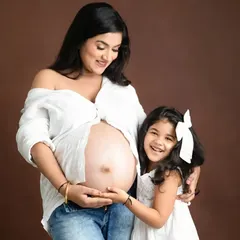 A joyful family moment captured against a warm brown backdrop. The daughter's happy expression as she touches her mother's baby bump makes this a precious memory.