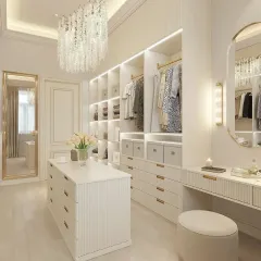 A bright and elegant walk-in closet in a soft cream palette. The design includes a central storage island, a vanity with a lighted mirror, and extensive open and closed wardrobe space with integrated LED lighting.