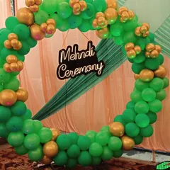 Here is a simple yet stylish ring backdrop we created for a Mehndi ceremony. The full circle is covered in a lush garland of green and gold balloons, which looks great in photos. A custom 'Mehndi Ceremony' sign is placed in the center, and we can adjust the colors to match your theme perfectly.