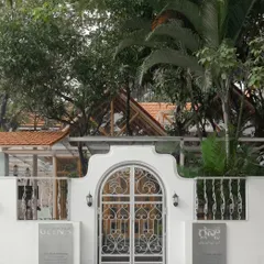 The white boundary wall and arched gate create an inviting entry point, contrasting with the lush greenery and the wooden structure of the cafe visible behind it.