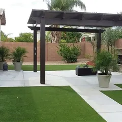 A simple yet elegant dark wood pergola installed over a concrete patio. This structure provides partial shade and a frame for future climbing plants, effectively defining the outdoor living space next to the lawn.