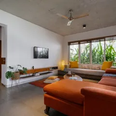 In this living room, a large window with a built-in bay seat is framed by lush greenery instead of traditional curtains. The vibrant orange sectional sofa adds a bold splash of color against the neutral palette of white walls and a concrete ceiling.