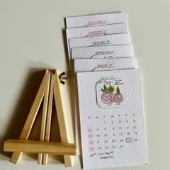 Here are the calendar pages for the second half of the year, stacked next to the wooden easel they come with. It's a complete, ready-to-display set.