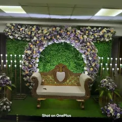 A lush green backdrop for a Half-Saree function in JP Nagar. The artificial green wall is adorned with a thick arch of pastel purple and white flowers, creating a garden-like feel indoors.