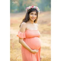 A beautiful, direct portrait of the mom-to-be. Wearing a floral crown and an off-the-shoulder peach gown, her happy expression and radiant glow are the central focus of the image.
