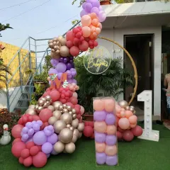 An outdoor first birthday setup on a grassy lawn. It features a gold ring backdrop with a neon sign, a large balloon arrangement in shades of pink and purple, and a light-up number '1'.